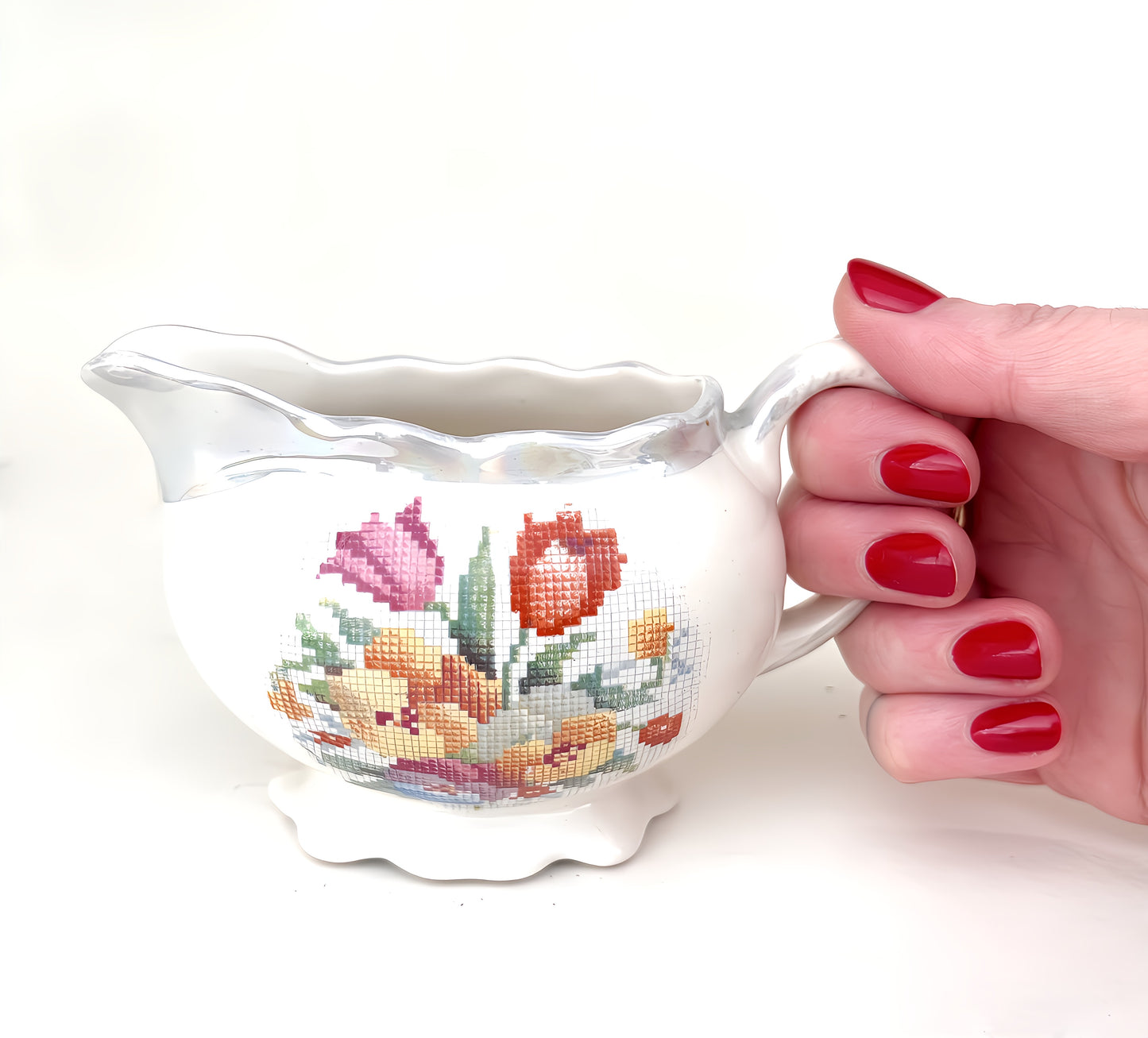 Close up of creamer with needlepoint design, held by handle. White background.