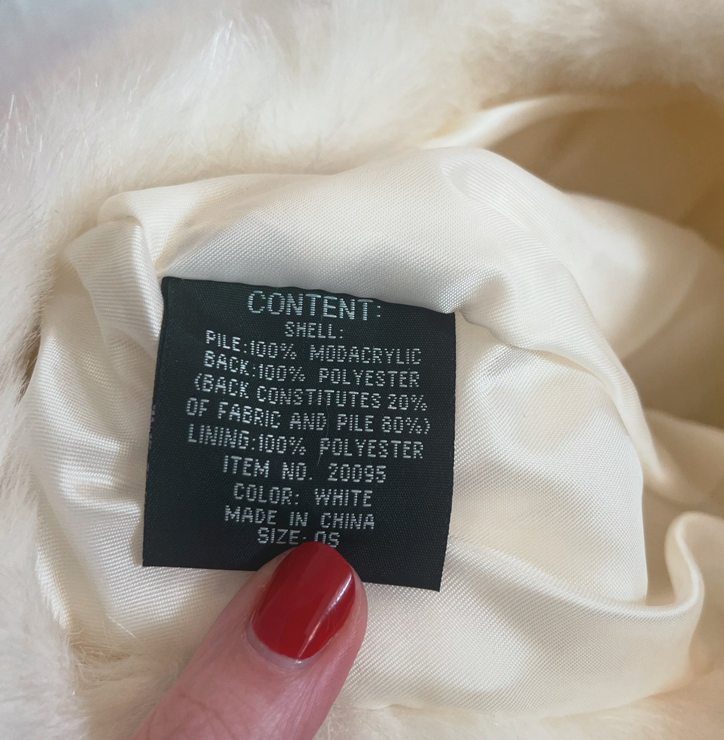 Close up shot of content tag which reads "CONTENT: shell: Pile 100% Modacrylic; Back: 100% Polyester (Back constitutes 20% of fabric and 80% of pile) Lining: 100% Polyester. Item No. 20095. Colour: White. Made in China. Size OS." White background.