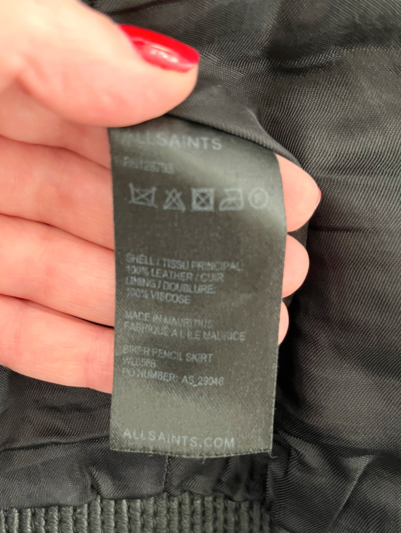 Close up of fabric tag which reads "All Saints. Shell/Tissu Principal 100% leather/Cuir. Lining/Doublure 100% Viscose. Made in Mauricius. Fabrique a le Maurice. Biker Pencil Skirt. Allsints.com"